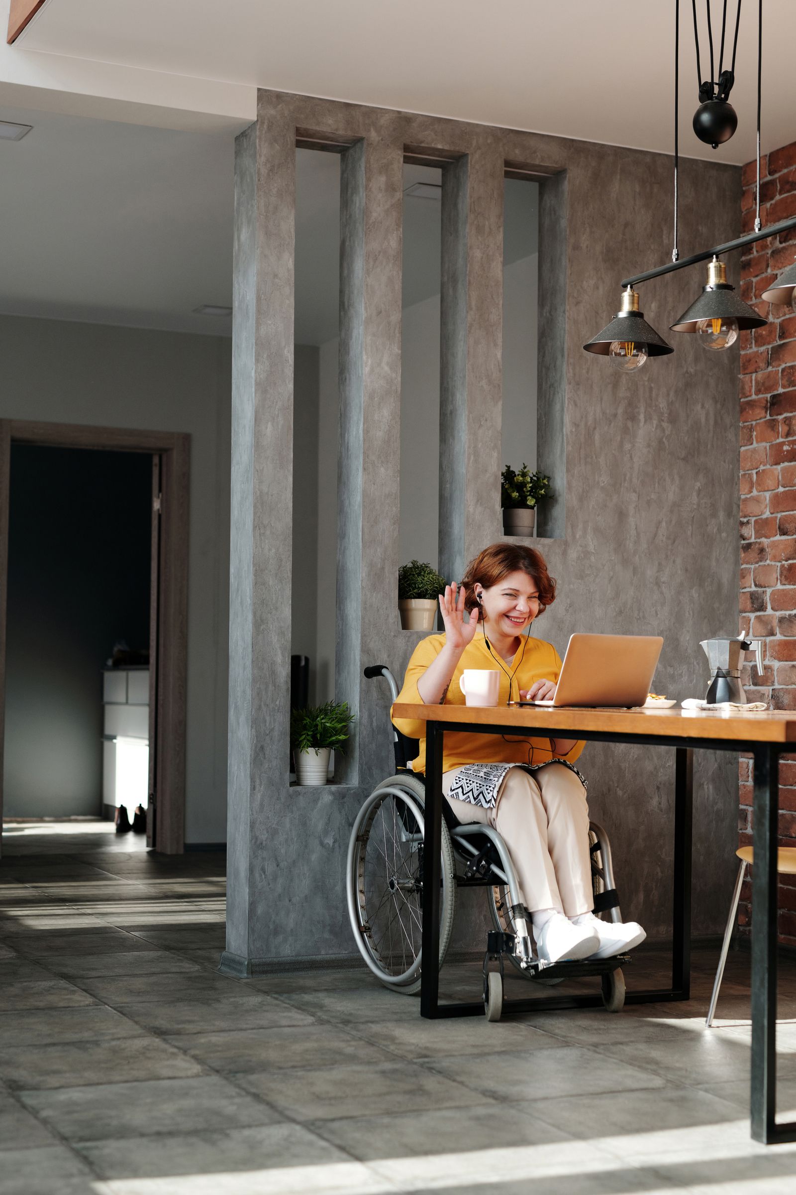 A smiling woman using a wheelchair waves during a video call at her kitchen table
